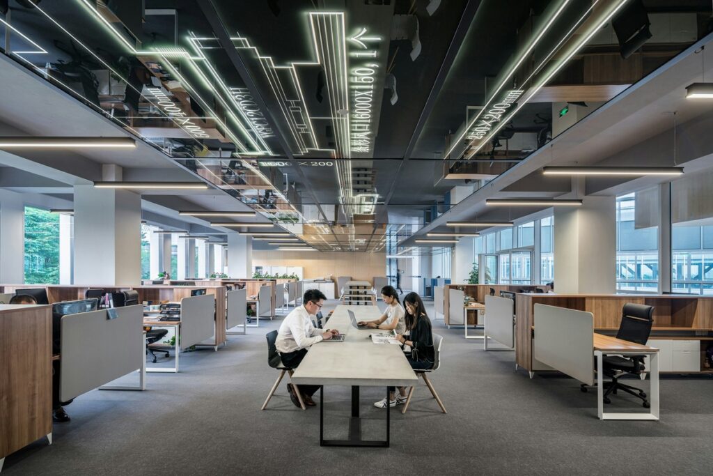 Modern open-plan office with three people working at a central table, surrounded by cubicles and illuminated ceiling designs